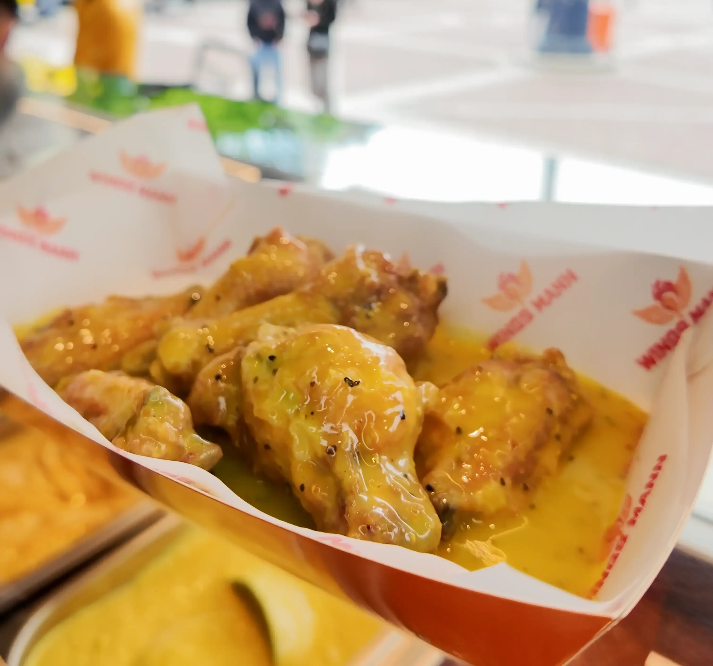 Ein Behälter mit knusprigen Hähnchenflügeln, die mit einer gelben Sauce überzogen sind. Der Behälter hat das WINGS MANN-Logo darauf.