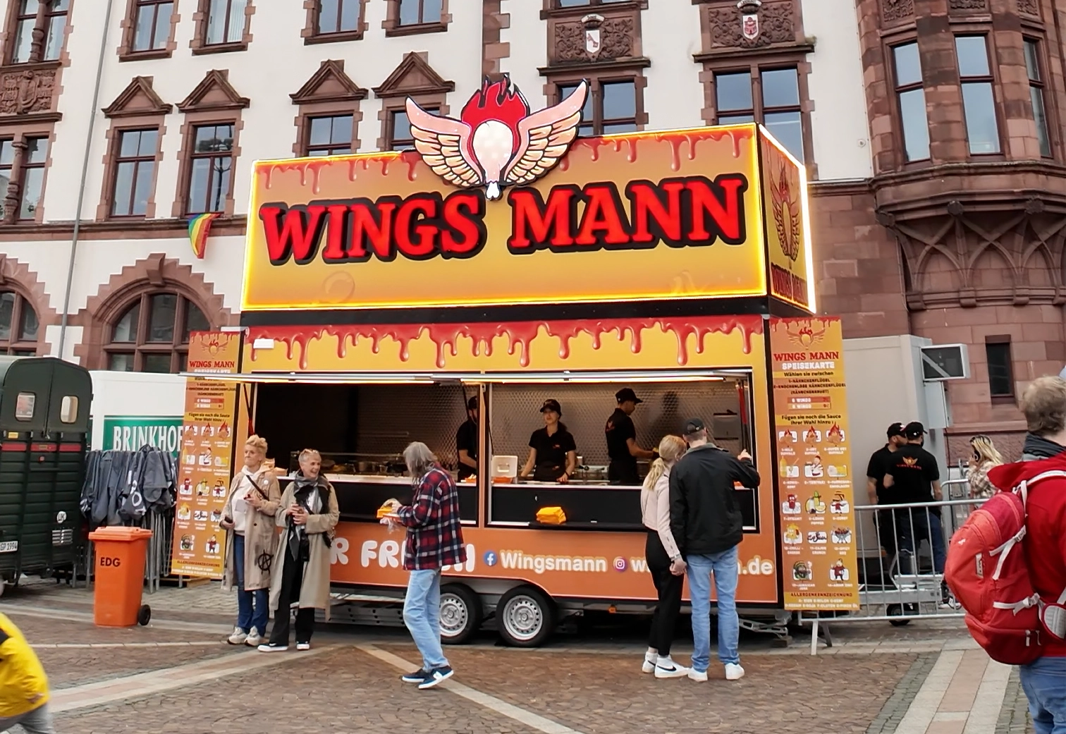 WINGS MANN Food-Truck, der frittierte Hähnchenflügel mit besonderen Saucen anbietet. Die auffällige Beschriftung in großen roten Buchstaben ist von flammenden Flügeln und tropfender Sauce umgeben. Die Vorderseite des Trucks ist geöffnet, um Kunden zu bedienen, und mehrere Personen stehen davor und bestellen Essen.